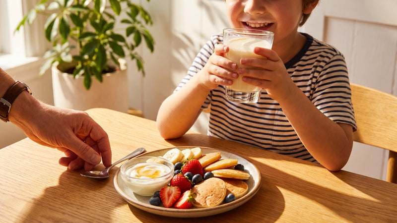 Lachendes Kind trinkt einen Vanille-Milchshake am Frühstückstisch mit bunten Pancakes und Beeren – wenn Kinder zu wenig essen