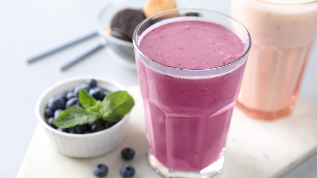 Glas mit cremigem Beeren-Smoothie neben einer Schale Blaubeeren mit Minze und einem hellen Milchshake im Hintergrund